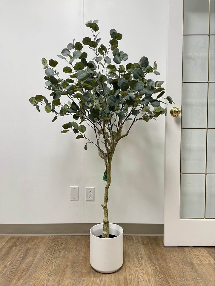 Artificial plant with broad, green leaves displayed in a modern, round white pot, placed next to a beige armchair, adding a natural touch to the indoor setting.