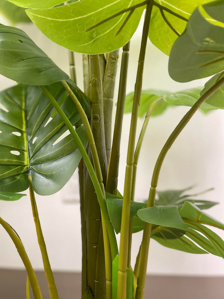 Close-up view of artificial monstera plant stems, featuring vibrant green shades and a natural texture. The image captures the details of the stems with a blurred foreground leaf, adding depth and realism to the plant&