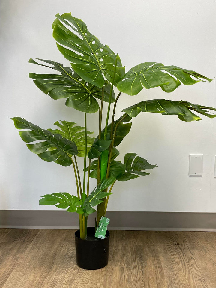 Artificial monstera plant in a black pot, placed on a wooden floor next to a modern white and gold-trimmed armchair. 
