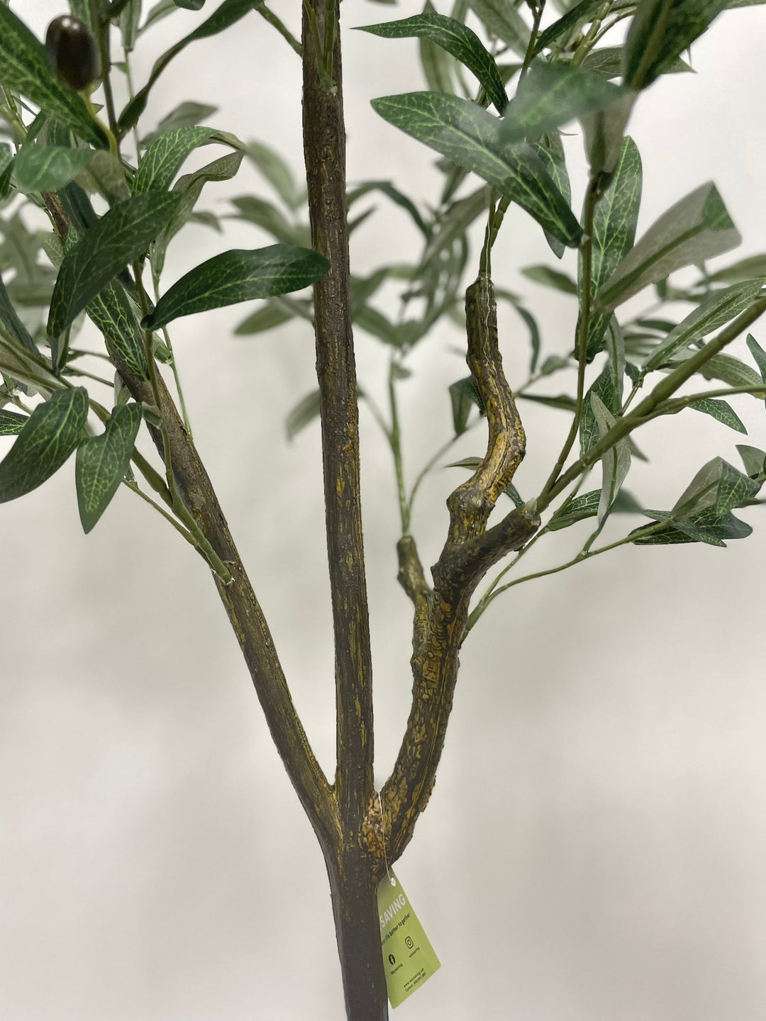Close-up of the detailed trunk and branches of an artificial olive tree with a textured, lifelike appearance. A green tag with the brand name "Waysaving" is attached to one of the branches. The tree features realistic bark textures and connected branches, making it a convincing decorative piece for indoor settings.