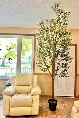 Tall artificial olive tree in a black pot placed beside a cream-colored recliner in a well-lit living room with large windows and wooden trim, showcasing a serene outdoor view.