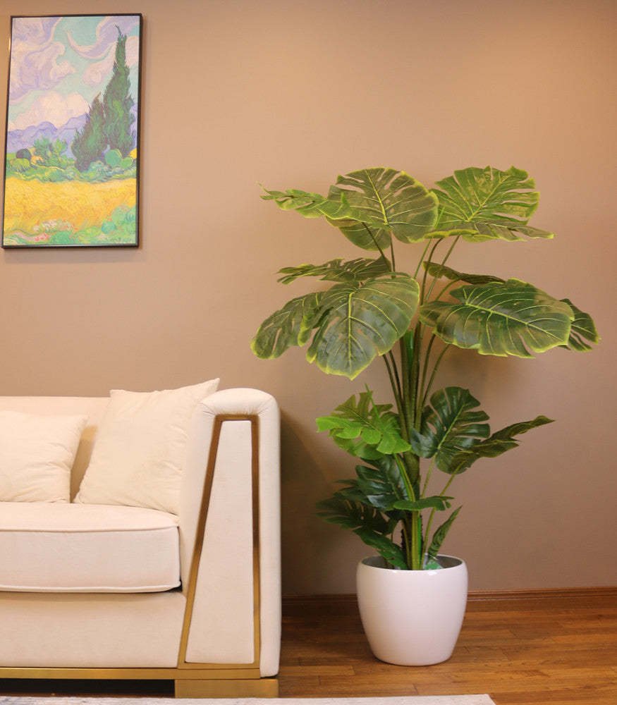 Artificial monstera plant in a white pot placed beside a modern beige sofa in a stylish living room. The lush green foliage adds a vibrant touch to the neutral-colored space, complemented by a colorful wall painting above the sofa, creating a cozy and inviting atmosphere