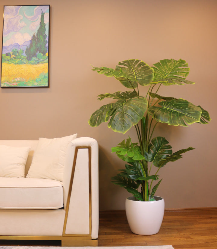 Artificial monstera plant in a white pot placed beside a modern beige sofa in a stylish living room. The lush green foliage adds a vibrant touch to the neutral-colored space, complemented by a colorful wall painting above the sofa, creating a cozy and inviting atmosphere