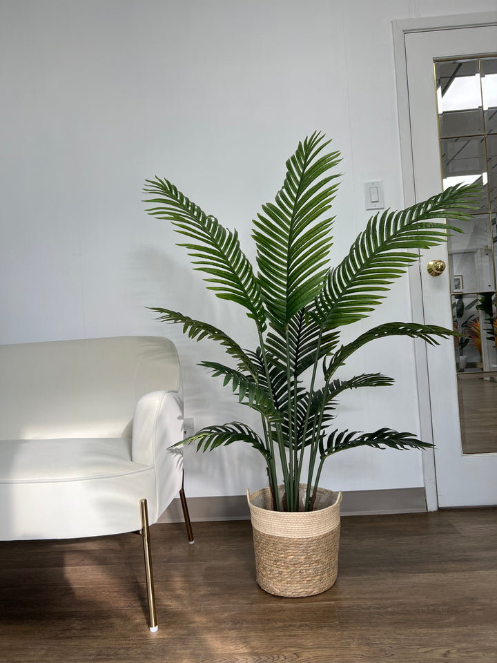 A large, artificial palm tree stands in a woven basket planter next to a cream-colored sofa. The setting is a cozy living room with warm lighting and neutral wall tones, creating a relaxed atmosphere.