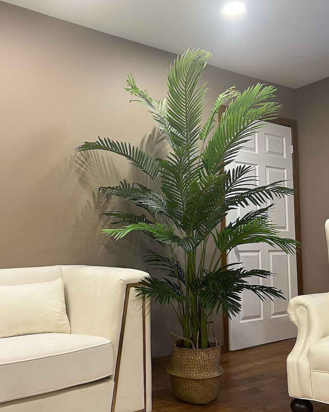 A large, artificial palm tree stands in a woven basket planter next to a cream-colored sofa. The setting is a cozy living room with warm lighting and neutral wall tones, creating a relaxed atmosphere.