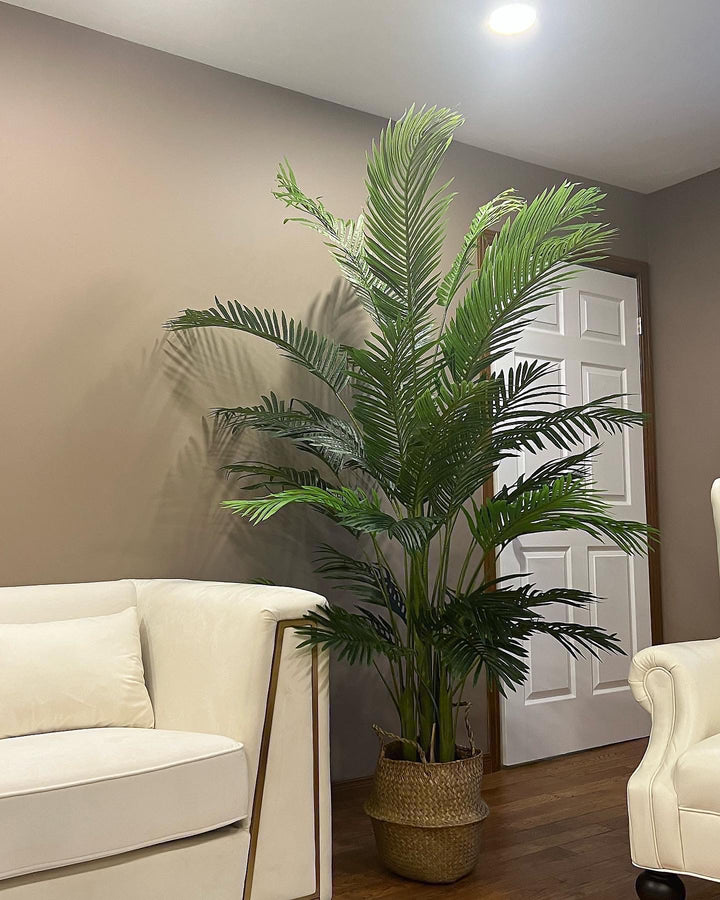 A large, artificial palm tree stands in a woven basket planter next to a cream-colored sofa. The setting is a cozy living room with warm lighting and neutral wall tones, creating a relaxed atmosphere.