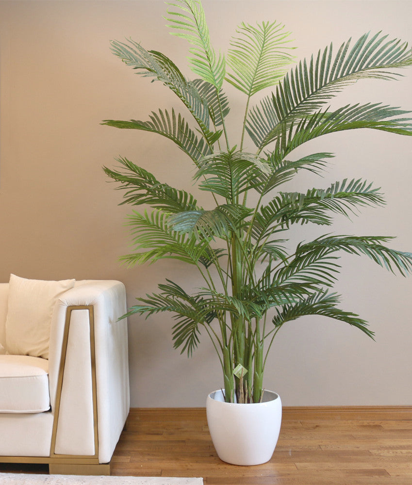 Artificial palm tree with green fronds, placed in a sleek white pot next to a white sofa, adding a natural touch to a modern, well-lit living room.