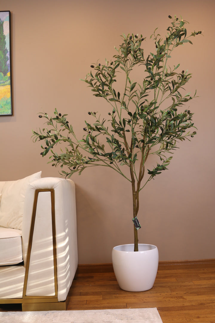 Artificial olive tree with slender branches and lifelike green leaves, adorned with small black olives, placed in a white ceramic pot beside a beige sofa with gold accents. The tree is set against a light brown wall, creating a warm and inviting interior scene