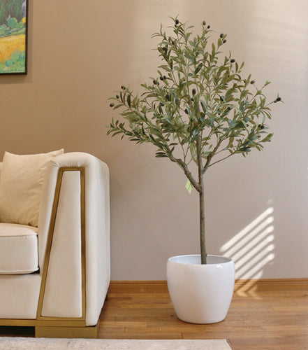 Elegant living room setup featuring a tall artificial olive tree in a white pot positioned next to a modern beige sofa with gold trim. The room has a warm, brown-toned wall adorned with a colorful landscape painting, creating a cozy and sophisticated atmosphere.