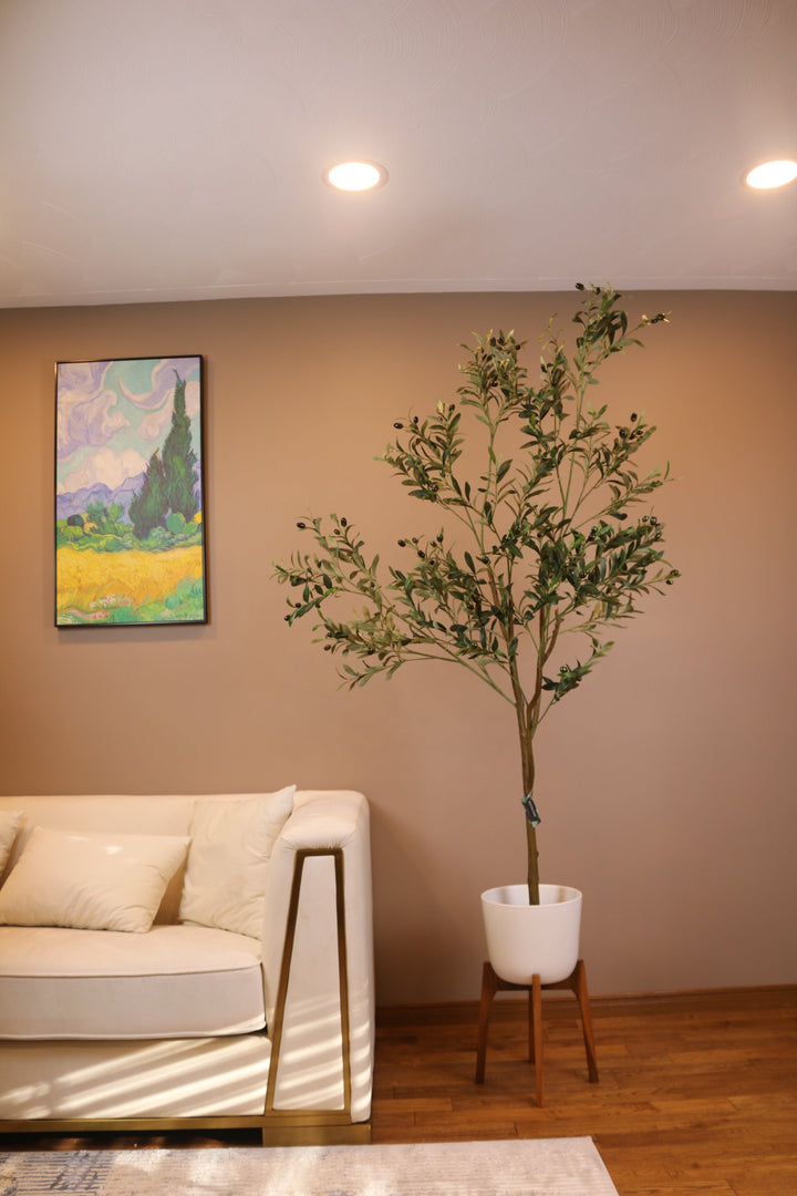 Elegant living room setup featuring a tall artificial olive tree in a white pot positioned next to a modern beige sofa with gold trim. The room has a warm, brown-toned wall adorned with a colorful landscape painting, creating a cozy and sophisticated atmosphere.