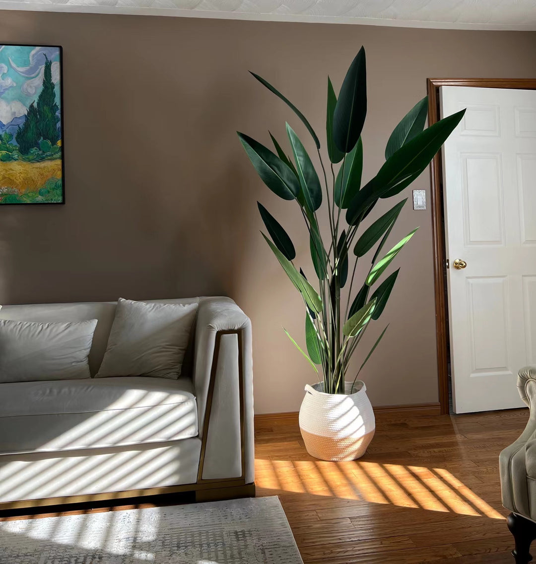 A cozy living room scene with a large artificial bird of paradise plant in a woven basket placed in the corner near a white door. The room is illuminated by soft natural light filtering through blinds, casting shadows on a modern beige sofa with matching cushions. A piece of colorful wall art hangs on the warm-toned wall above the sofa, complementing the wooden flooring and serene ambiance of the space.