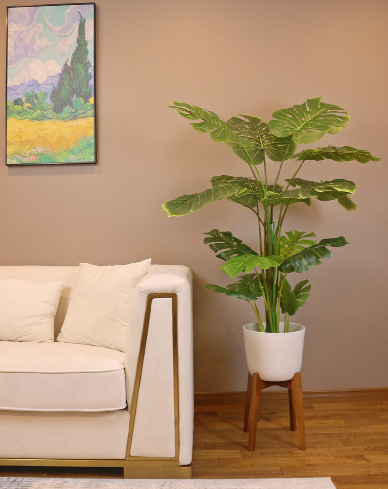 Artificial monstera plant in a white ceramic pot with a wooden stand, positioned next to a cream-colored armchair. The plant features large, realistic green leaves with natural splits, adding a tropical vibe to the indoor space. The setup is placed on a carpeted floor with subtle blue accents