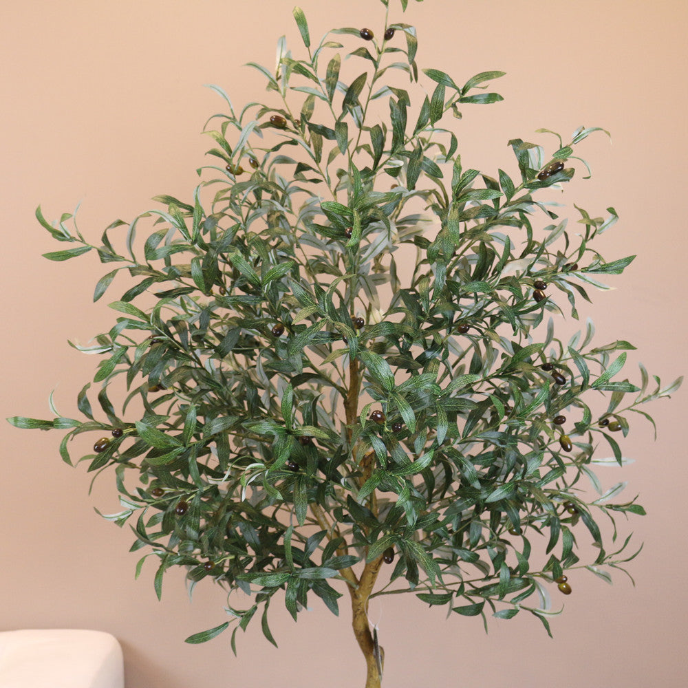 Artificial olive tree with dense green foliage and small black olives, standing against a neutral-colored wall. The leaves have a natural texture and veining, creating a realistic appearance.