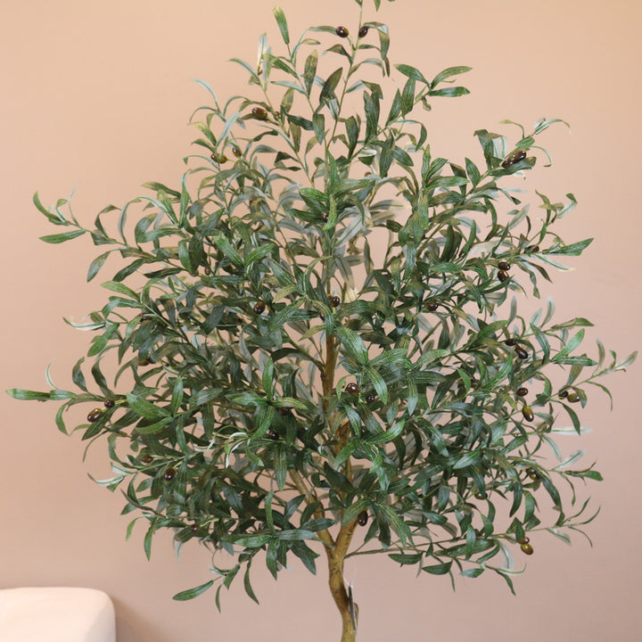 Artificial olive tree with dense green foliage and small black olives, standing against a neutral-colored wall. The leaves have a natural texture and veining, creating a realistic appearance.