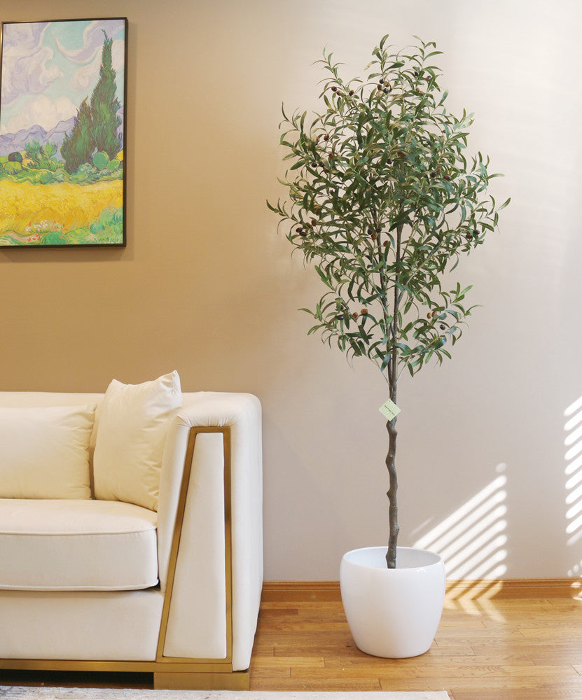 Artificial olive tree with slender branches and lifelike green leaves, adorned with small black olives, placed in a white ceramic pot beside a beige sofa with gold accents. The tree is set against a light brown wall, creating a warm and inviting interior scene.