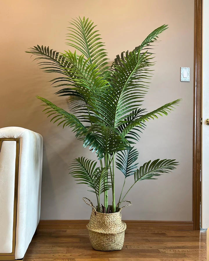 Artificial palm tree with green fronds, placed in a sleek white pot next to a white sofa, adding a natural touch to a modern, well-lit living room.