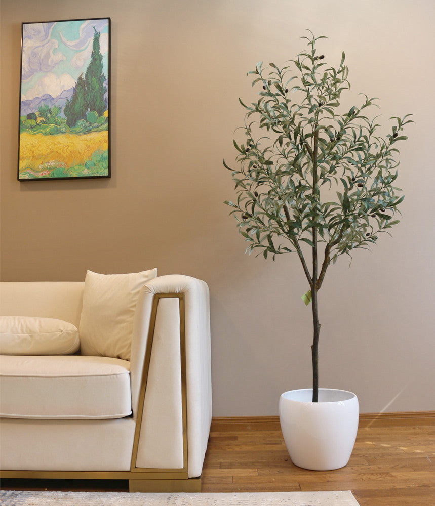 Artificial olive tree with slender branches and lifelike green leaves, adorned with small black olives, placed in a white ceramic pot beside a beige sofa with gold accents. The tree is set against a light brown wall, creating a warm and inviting interior scene