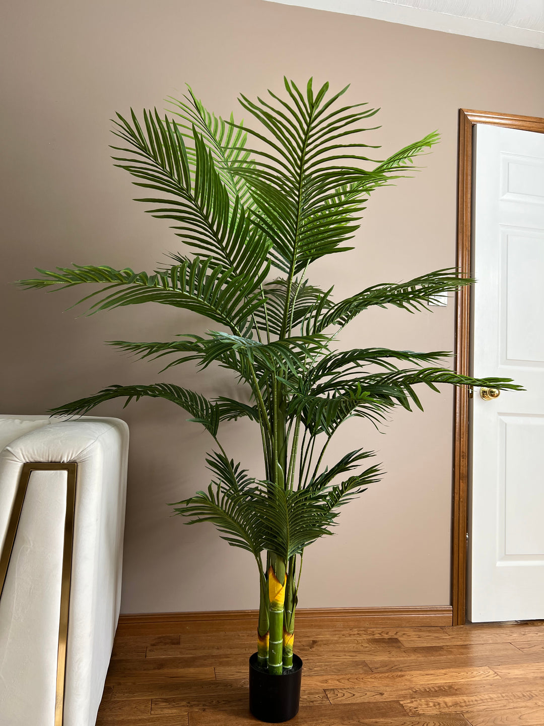A tall artificial palm tree with vibrant green leaves, placed in a simple black pot on a wooden floor, next to a modern white couch and a neutral-toned wall. The tree adds a lush, tropical feel to the space.